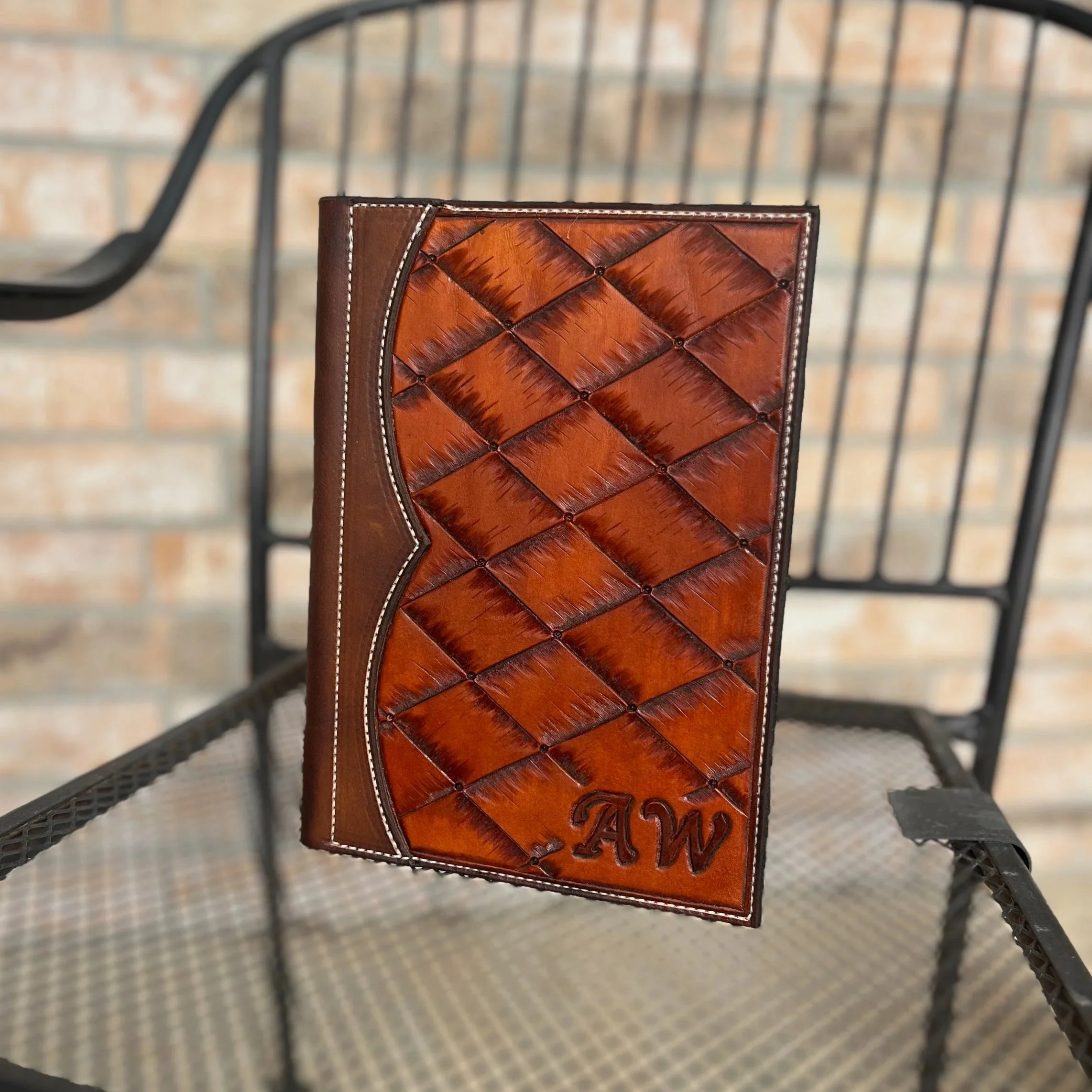 Close-up of a brown leather purse with a woven pattern, stitched edges, and a cut-out section revealing a logo or text, placed on a black metal outdoor chair with a woven seat, against a background of a brick wall.