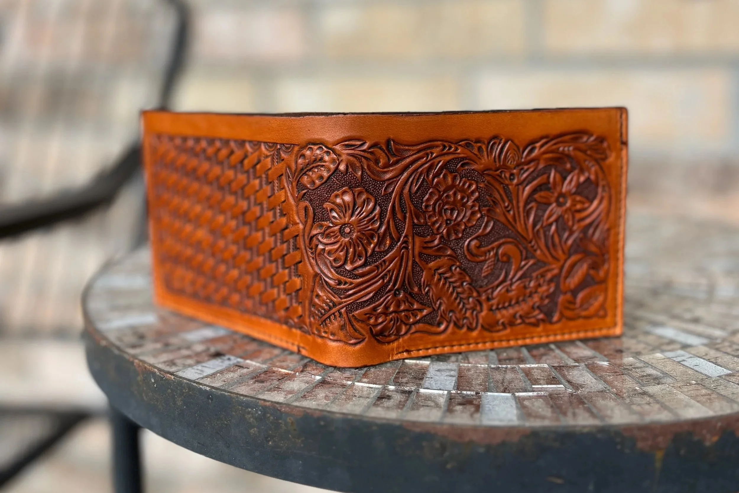Close-up of a brown leather wallet with intricate carved floral and scroll designs, resting on a textured surface.