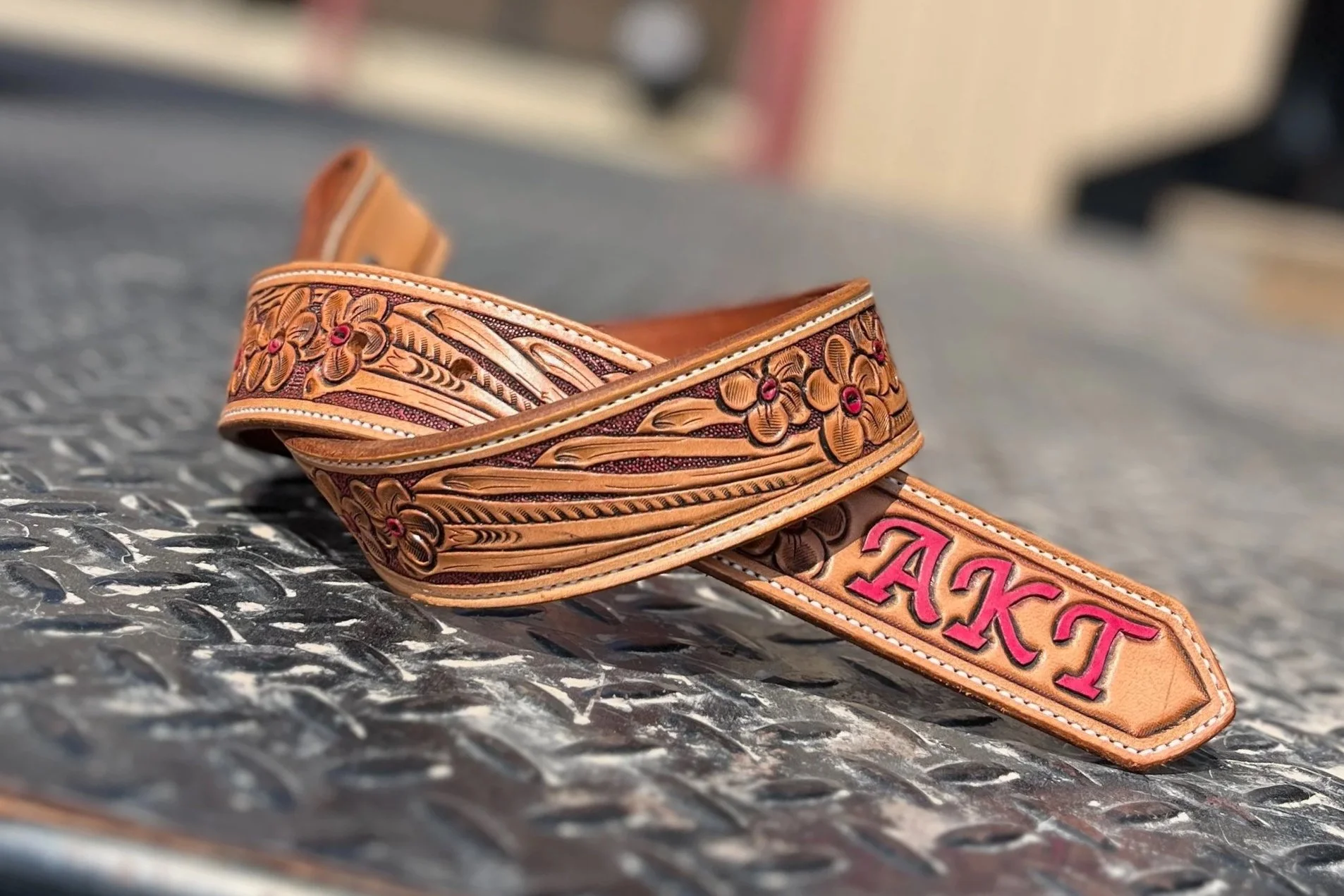 Close-up of a decorative leather belt with floral patterns and the word 'TAKIS' embroidered in pink, resting on a textured metallic surface.