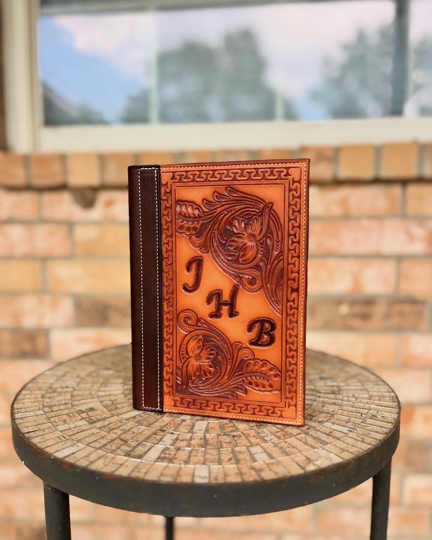 A leather-bound menu with intricate floral and scroll designs, and the word 'JH' embossed on the front, placed on a round mosaic-tiled table.