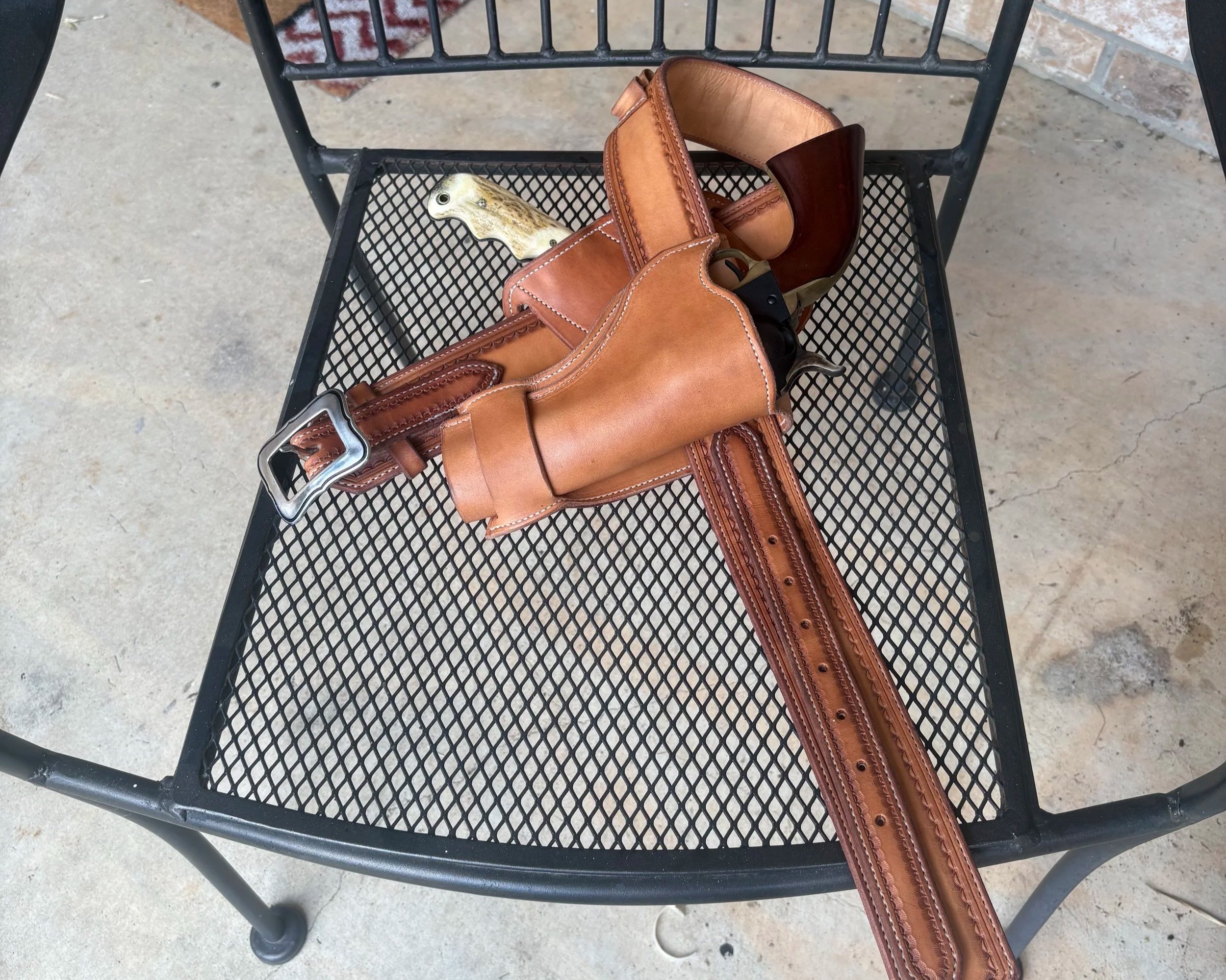 A black metal outdoor table with a mesh surface holding a brown leather bag and a beige gun with a decorative grip, with a concrete patio and brick wall in the background.