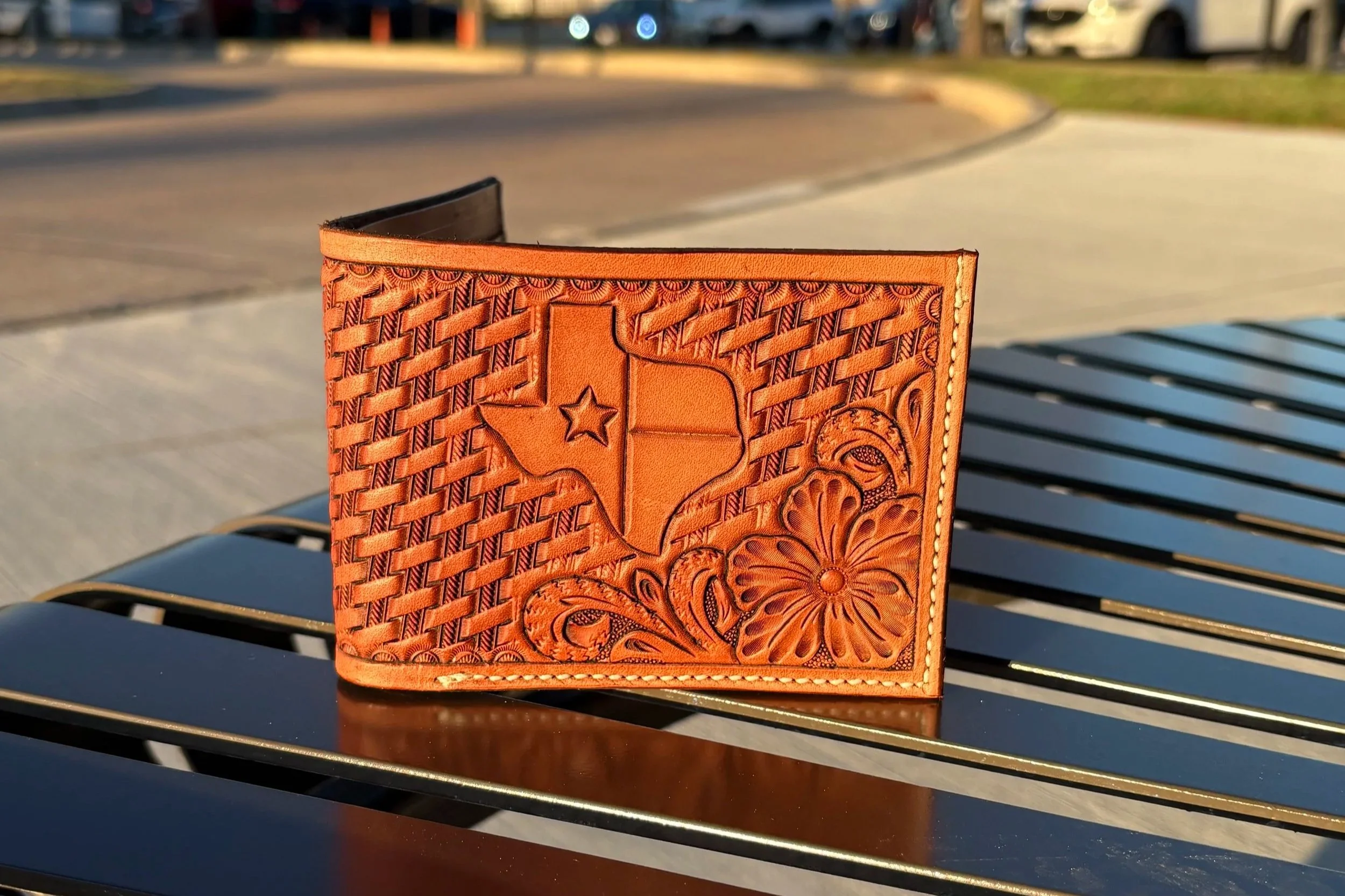 Close-up of an orange leather wallet with intricate floral and geometric embossed designs, sitting on a black metal outdoor bench.