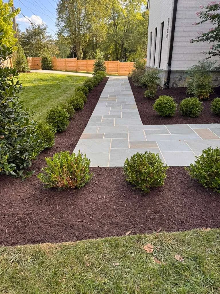 A paved stone walkway leading from a grassy lawn to a wooden fence, flanked by neatly trimmed bushes and mulch beds on either side, next to a white house with black downspouts.