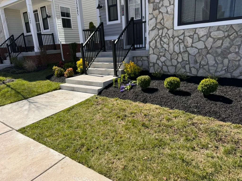 A modern house with a light-colored stone facade and a small front yard. There are concrete steps with black metal railings leading up to the front door. The yard has a well-maintained lawn and a flower bed with small bushes and some purple and yello