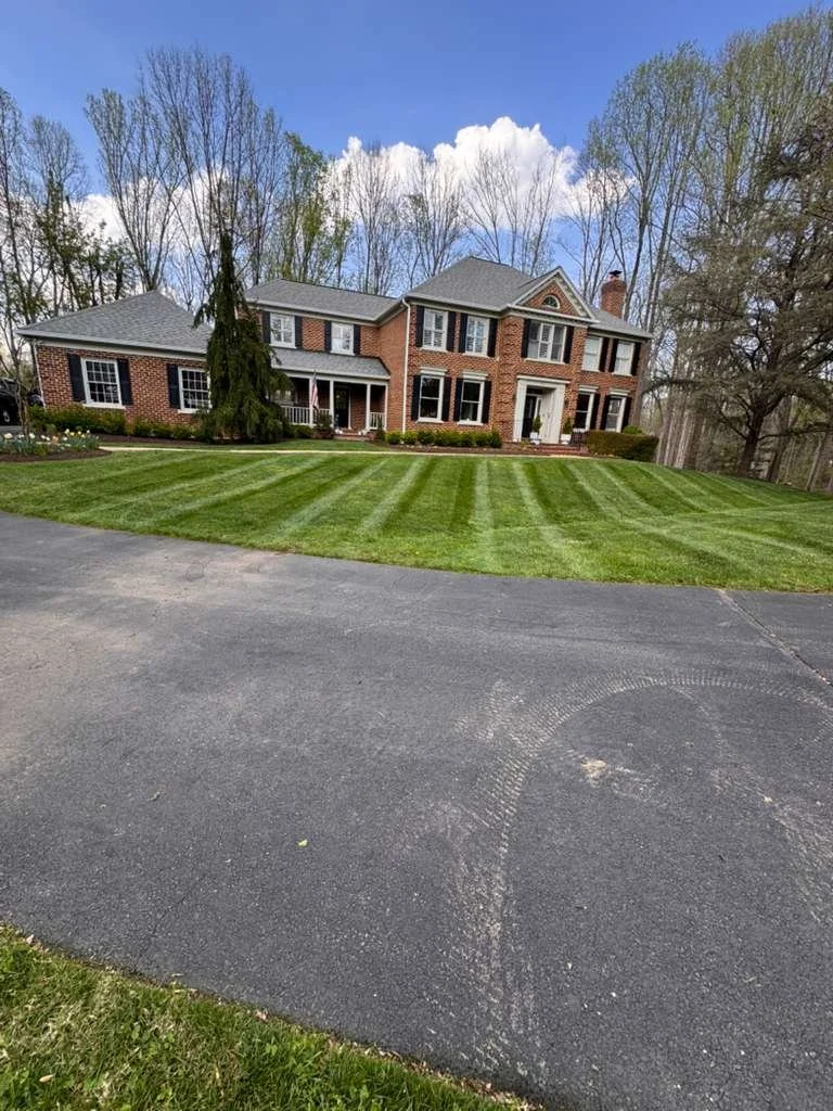 Large brick house with a well-maintained lawn, surrounded by tall trees, under a partly cloudy sky.