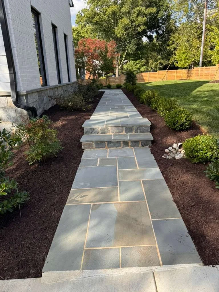 A newly paved stone walkway leads to a small set of stairs next to a house with a white brick exterior. The walkway is bordered by landscaped garden beds with shrubs and mulch. The background features a lush green lawn, trees, and a wooden fence.