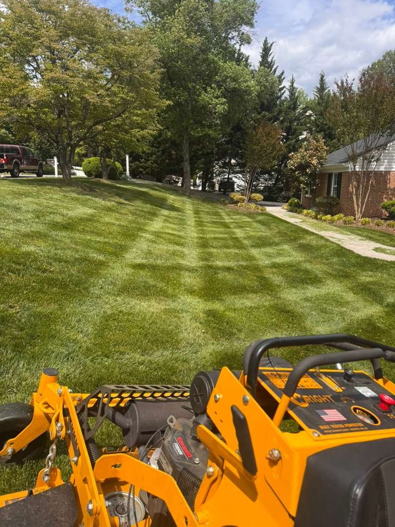 View from a lawn mower or landscaping equipment showing a neatly mowed grassy yard with hit stripes, trees, a sidewalk, a brick house, and parked vehicles in the background.