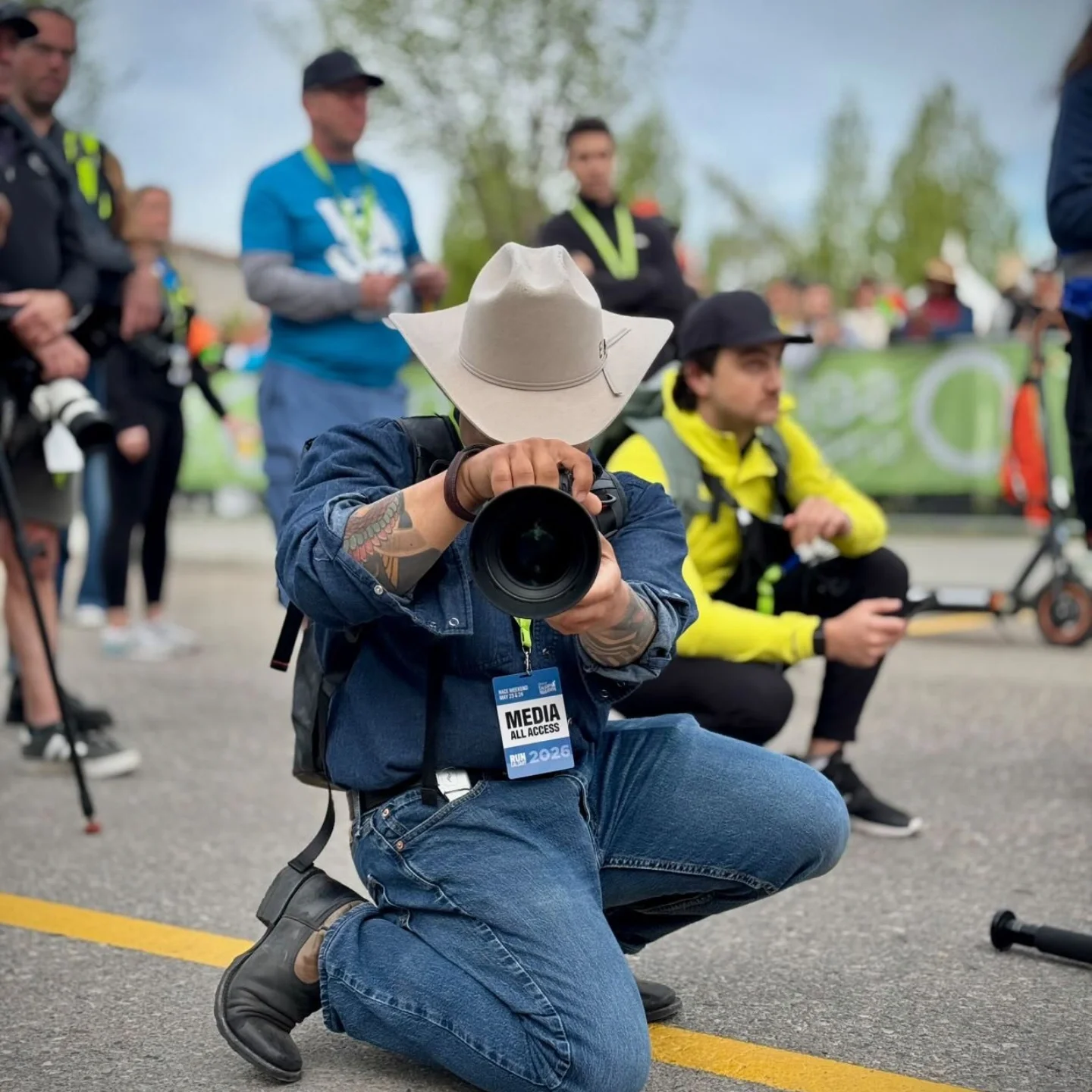 A photographer wearing a beige wide-brimmed hat and a media badge is kneeling on the ground, aiming a camera. Several people, some with cameras and media badges, are standing or sitting around him at a public outdoor event with trees and tents in the background.