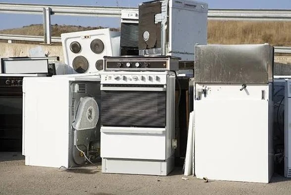 A pile of discarded white appliances, including stoves, refrigerators, and microwave ovens, outdoors on a concrete surface with a highway and landscape in the background.