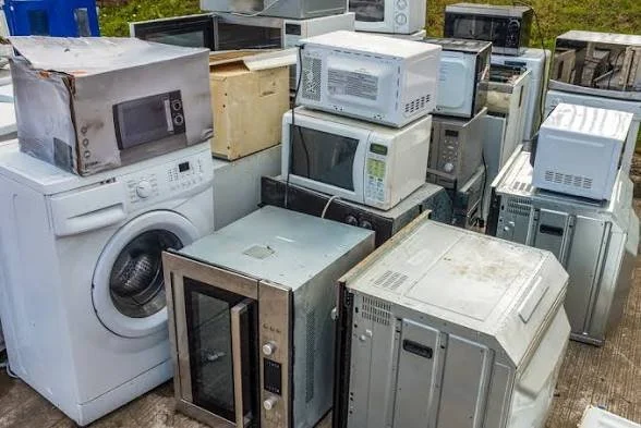 A collection of discarded microwaves, washing machines, and other appliances outdoors on concrete.