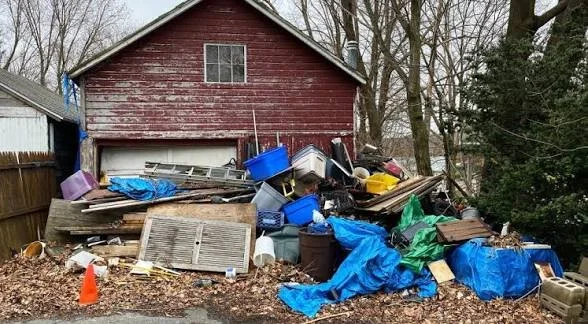 Piles of debris, trash, and discarded items in front of a red wooden house, including plastic bins, wooden panels, and blue tarps.