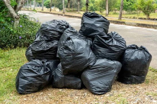 Stack of black garbage bags filled with trash on the side of the street next to a grassy area with trees and bushes.