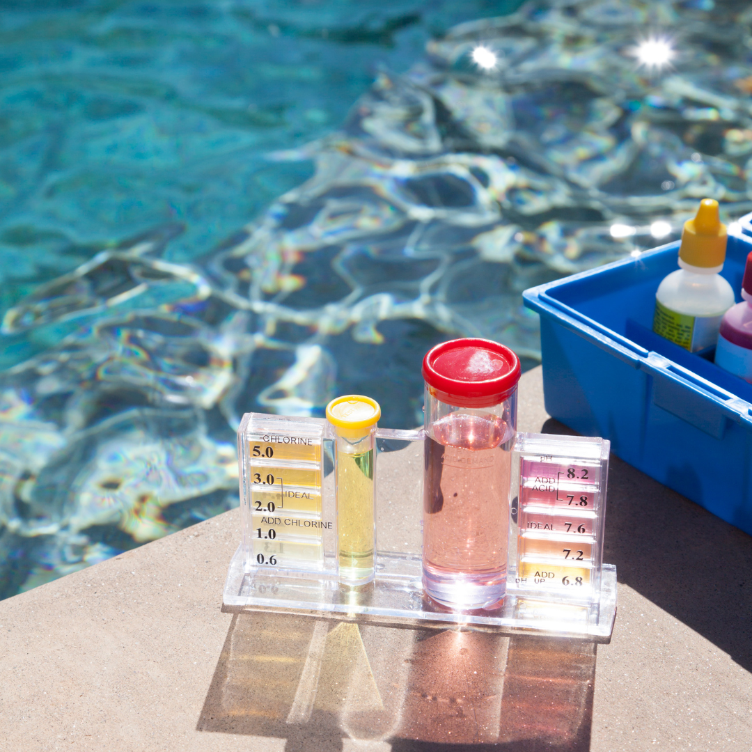 Poolside water testing kit with test tubes showing chlorine and pH levels, and bottles of testing chemicals in a blue container beside a swimming pool.