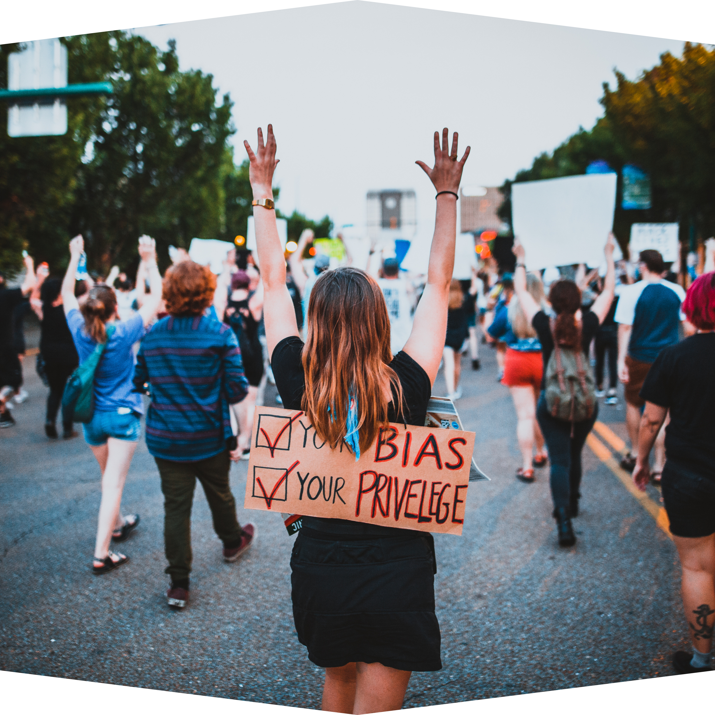 A view of a protest from behind with a white woman wearing a sign that reads "Check your bias. Check your privilege."