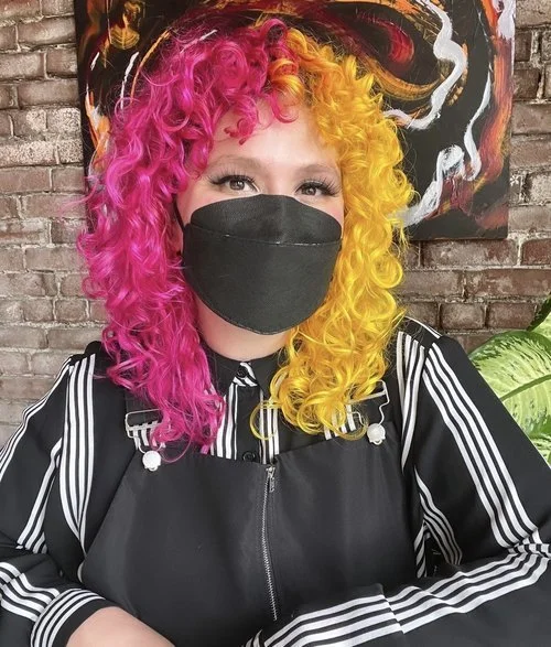 A photo of Noor. They are a light skinned Lebanese, femme presenting person with pink and yellow hair. They are wearing a KN-95 mask.