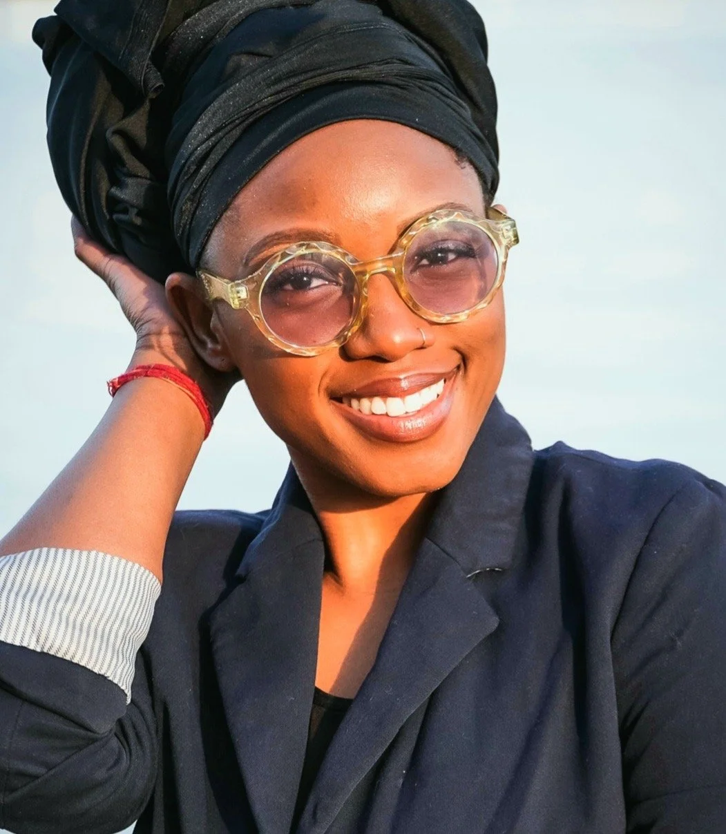 A photo of ave': a black, femme presenting person with deep brown skin, glasses, and a black turban. They are smiling.