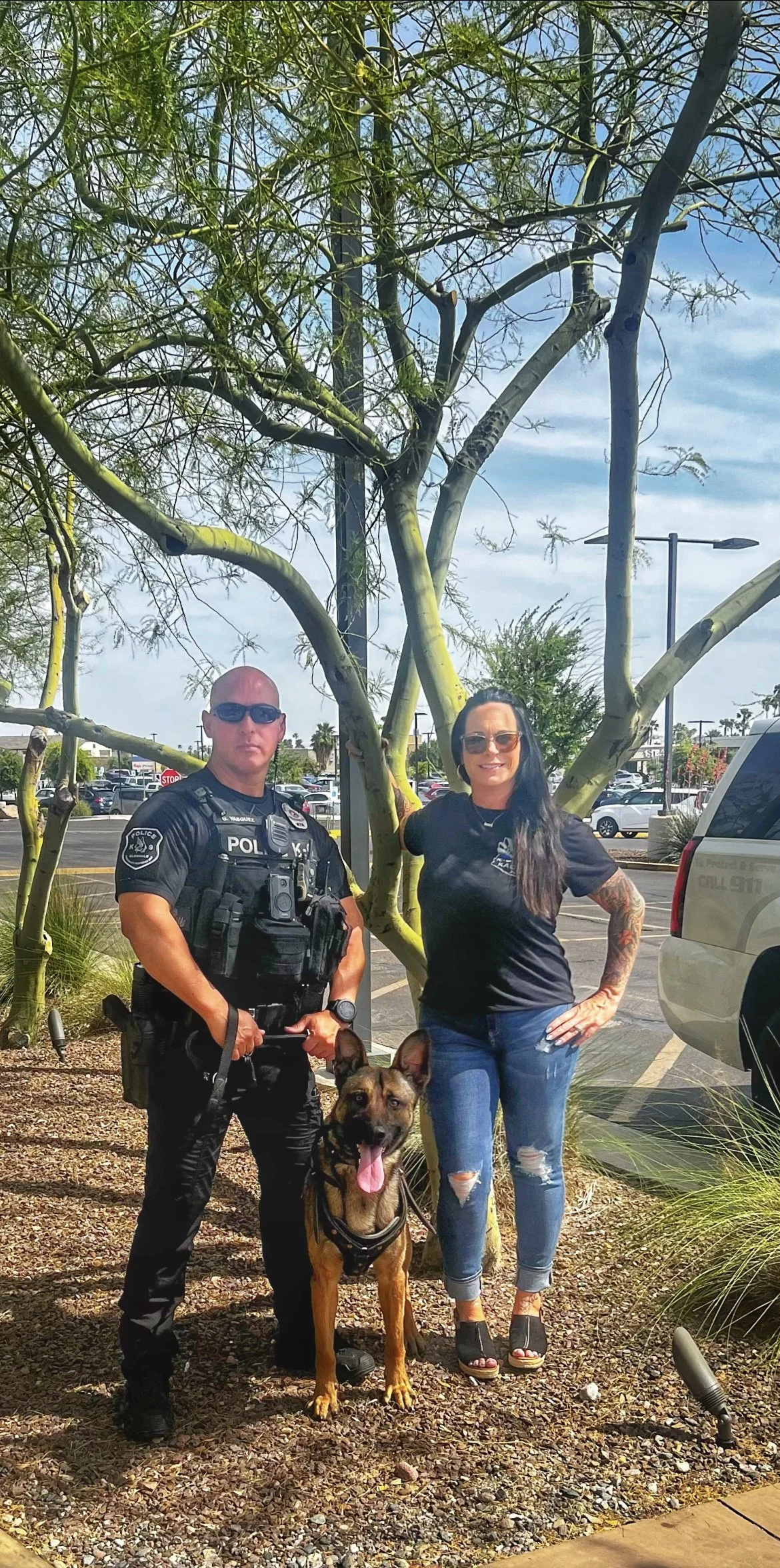 A police officer in tactical gear, a woman with tattoos and sunglasses, and a police service dog with a harness are posing in a parking lot with trees and parked cars in the background.