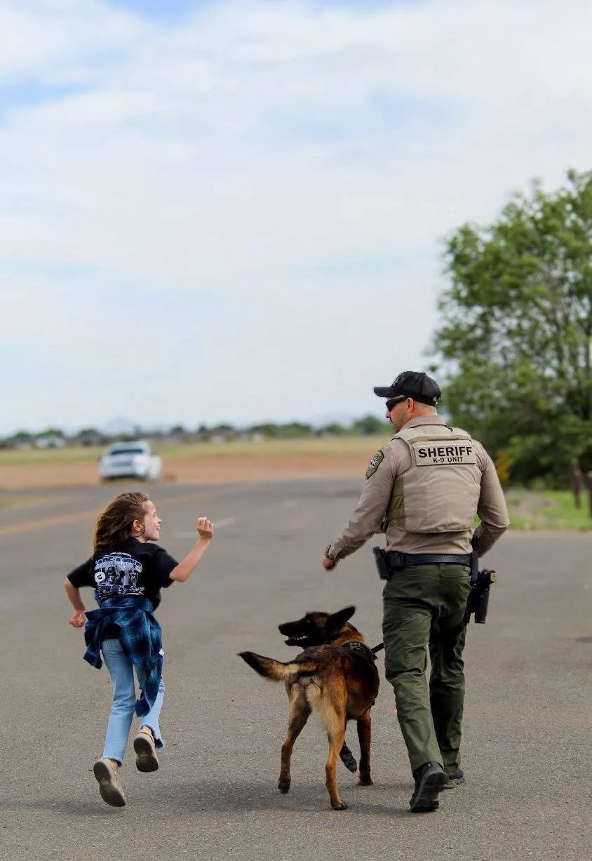 A girl running joyfully toward a sheriff's deputy and police dog in an open area with a dirt road and a tree in the background.