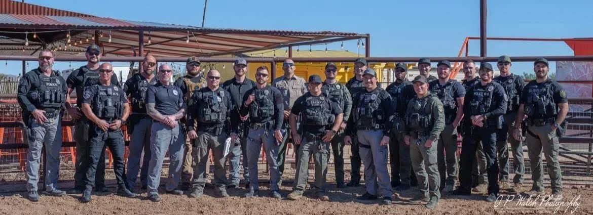 Group of police officers in uniform posing outdoors in front of a metal fence and colorful structures.