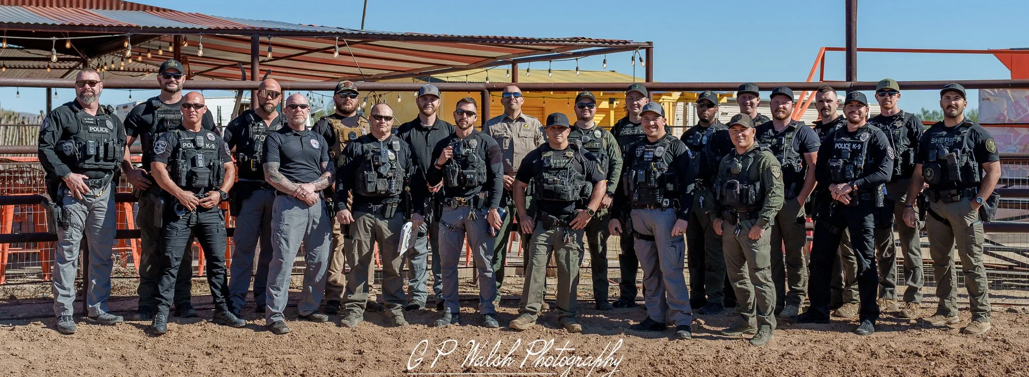 A group of uniformed police officers, sheriff deputies, and law enforcement personnel standing outdoors in front of a colorful structure with a clear blue sky.