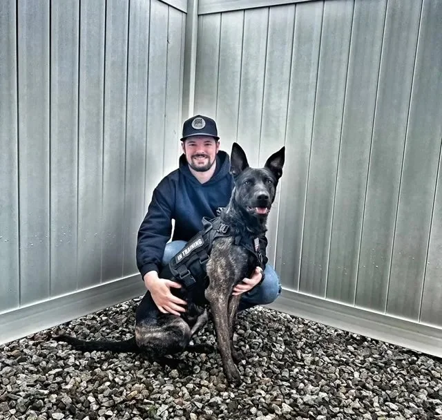 A man with a beard wearing a black baseball cap and navy blue hoodie is kneeling next to a black and brindle Belgian Malinois dog in a small outdoor enclosed space with metallic walls and gravel ground.