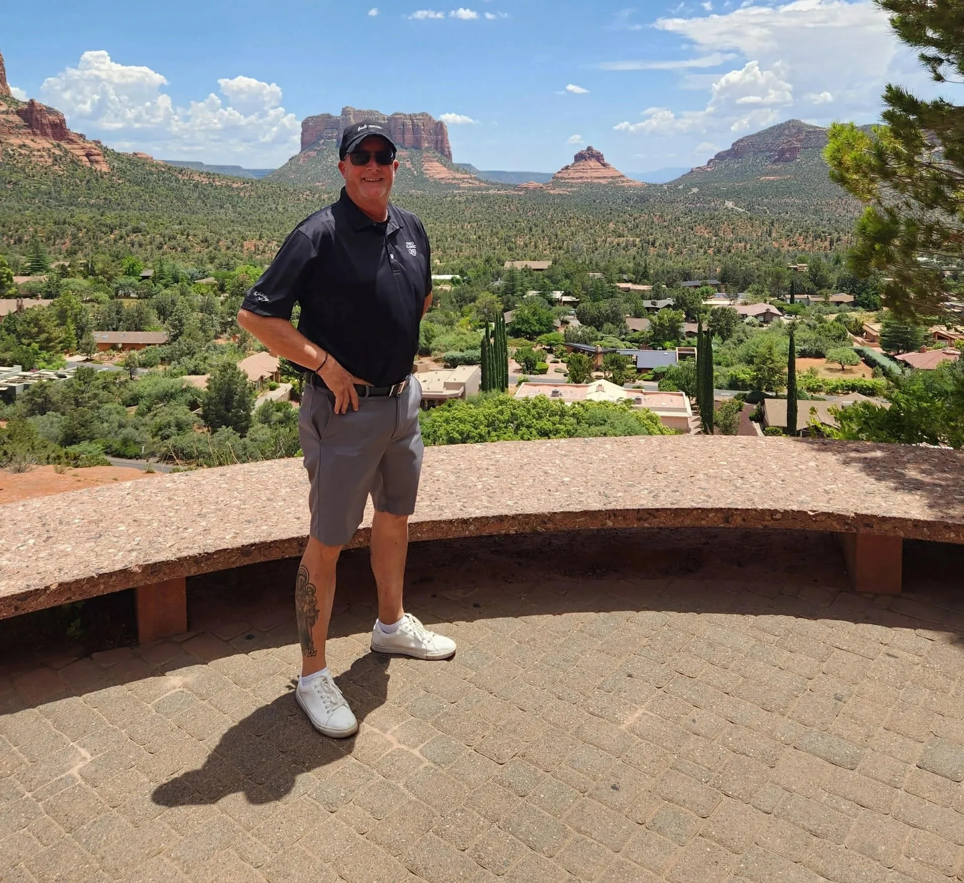 A man standing outdoors in front of a desert landscape with red rock formations and green vegetation, wearing a black hat, sunglasses, black polo shirt, gray shorts, and white sneakers.