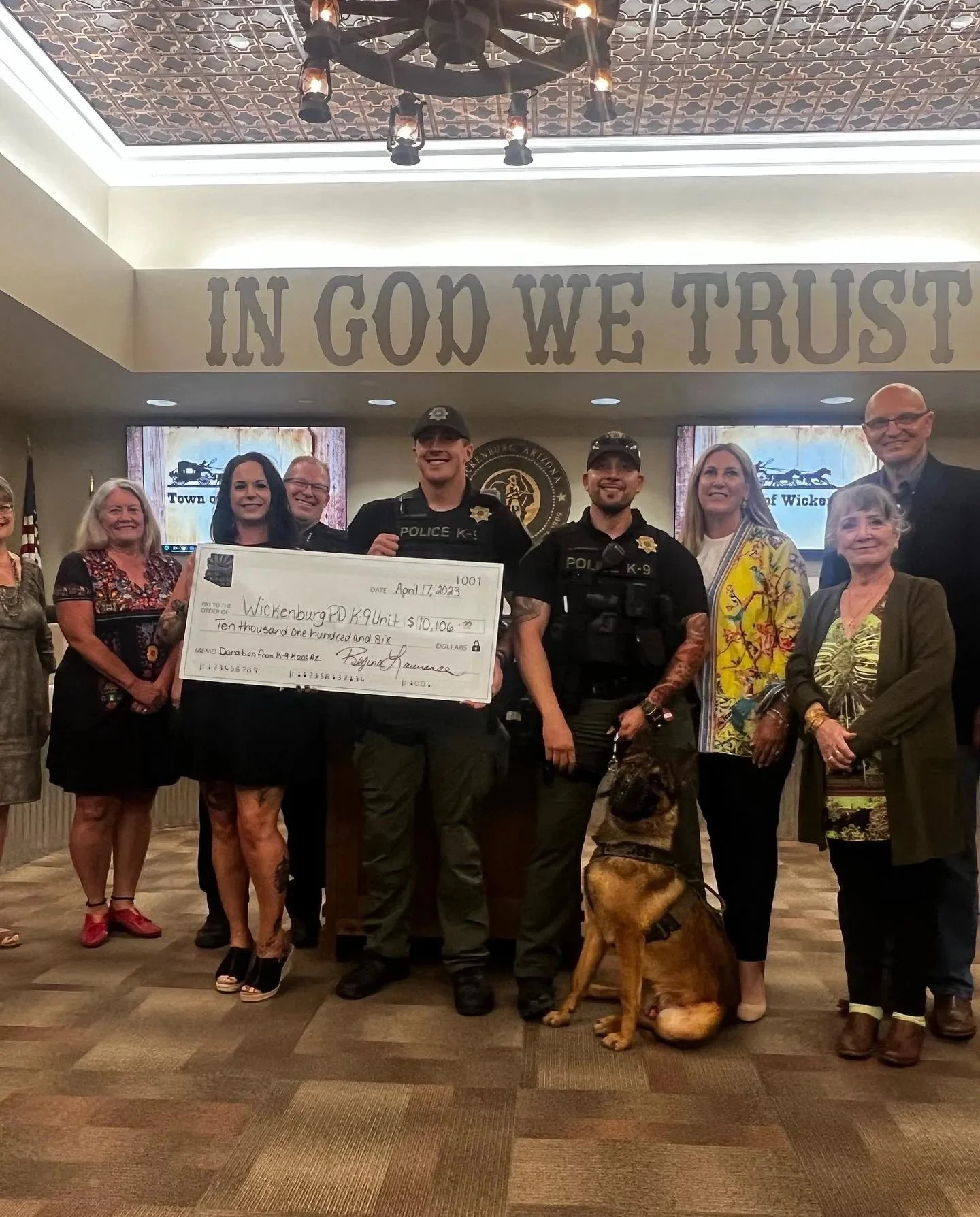 Group of people, including police officers, standing together in a room, holding a large check for $10,106.00, with a police dog sitting in front of them. Behind them, a sign reads 'IN GOD WE TRUST' and there are screens displaying a town seal.