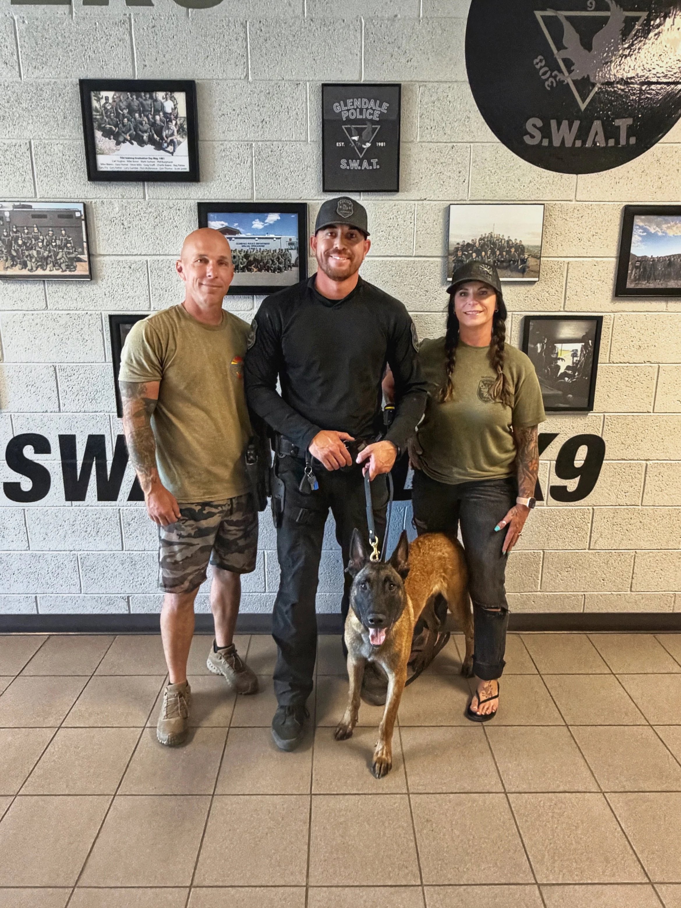 Three people standing in front of a wall with framed photographs and police insignia, with a police dog in front of them. The man in the middle is a police officer in uniform, smiling, with a Belgian Malinois dog at his feet. The other man has a tattoo sleeve on his left arm and is wearing a tan t-shirt and camouflage shorts. The woman has braided hair, wearing a camouflage t-shirt and black boots. The wall behind them features various police-related images and badges.