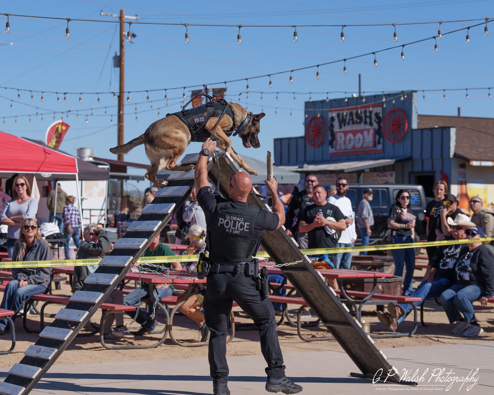 A police dog is jumping over an obstacle at an outdoor event with spectators watching. The event appears to be a demonstration or competition. The scene includes people sitting and standing, some smiling, and a police officer managing the dog. There 