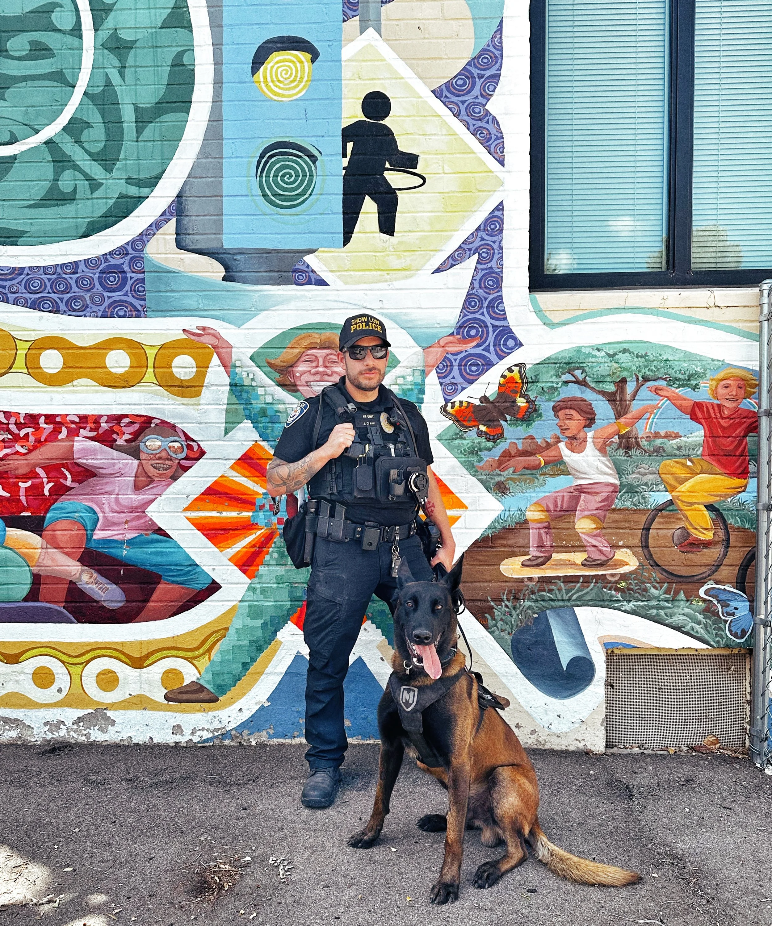 A police officer in uniform with a tactical vest and sunglasses standing in front of a colorful mural with children, a butterfly, and other artistic elements, holding a large German Shepherd dog on a leash.