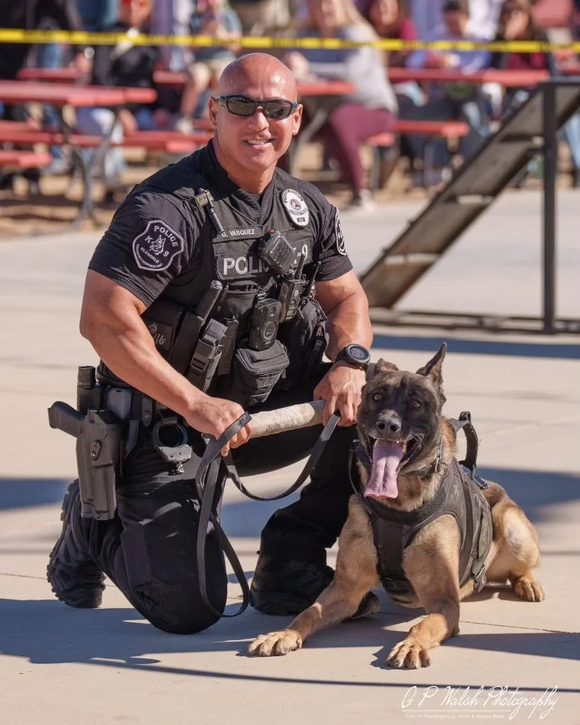 A smiling police officer kneeling next to a German Shepherd police dog in uniform during an outdoor event, with spectators in the background.