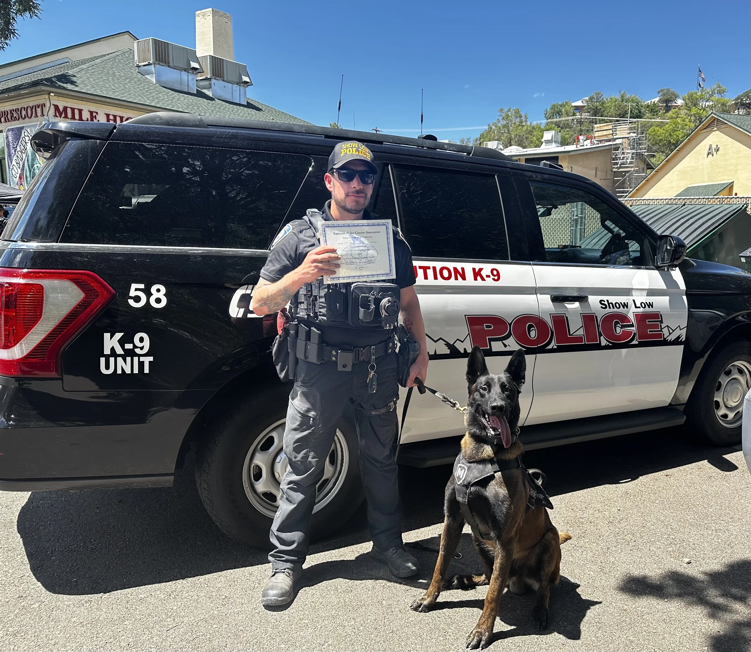 A police officer holding a certificate standing next to a police vehicle with a German Shepherd police dog sitting in front of him. The vehicle has markings indicating it is a K-9 unit, and the officer is wearing sunglasses and a uniform. The backgro