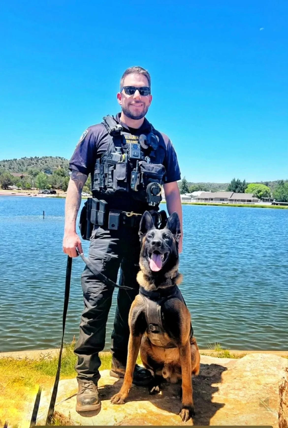 A police officer in uniform standing by a lake with a German Shepherd police dog, both looking at the camera under a clear blue sky.