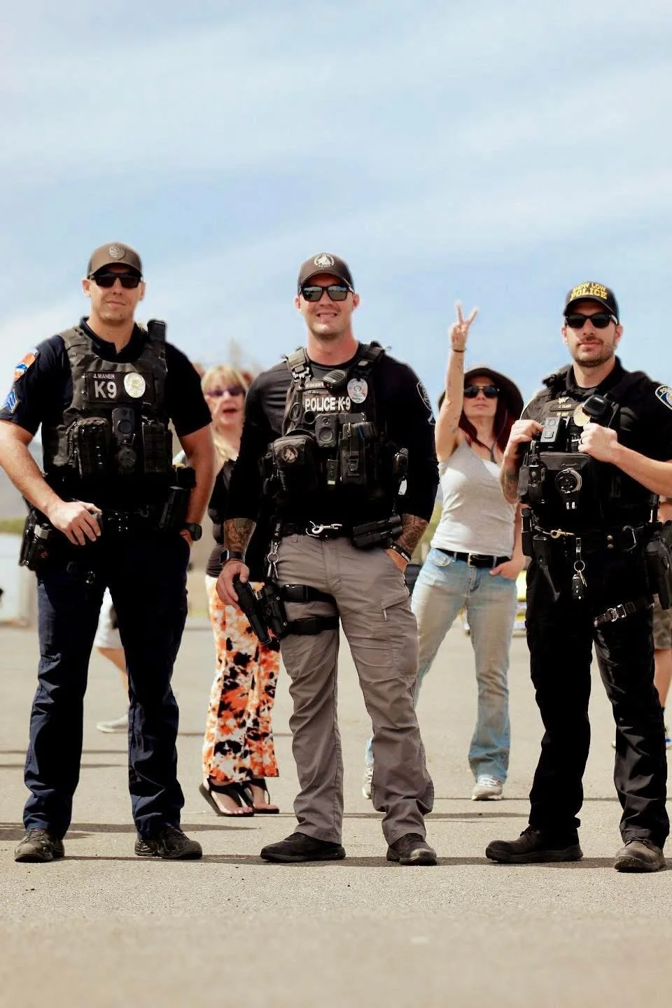 Three police officers in tactical gear standing outdoors, with three women in casual clothing and sunglasses behind them, one of whom is making a peace sign gesture.