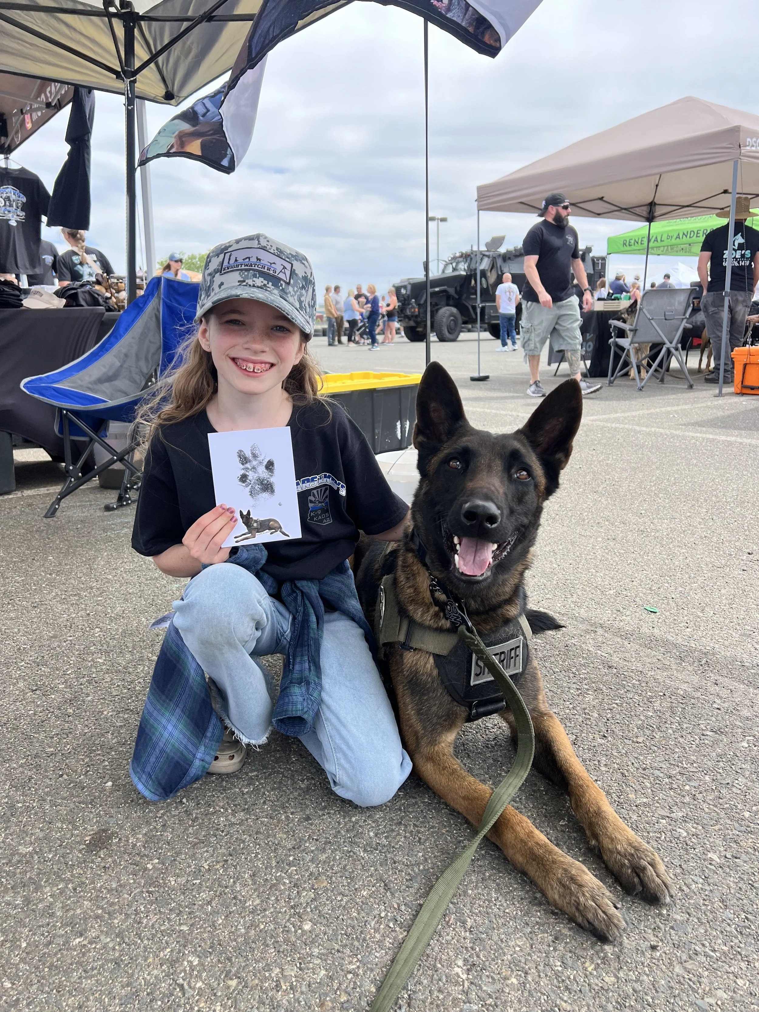 A young girl with long hair, wearing a camouflage cap, smiling and holding a drawing of a dog paw print. She is sitting on the ground next to a police dog with a harness labeled "SHERIFF." The scene is outdoors at a public event with tents, people, a
