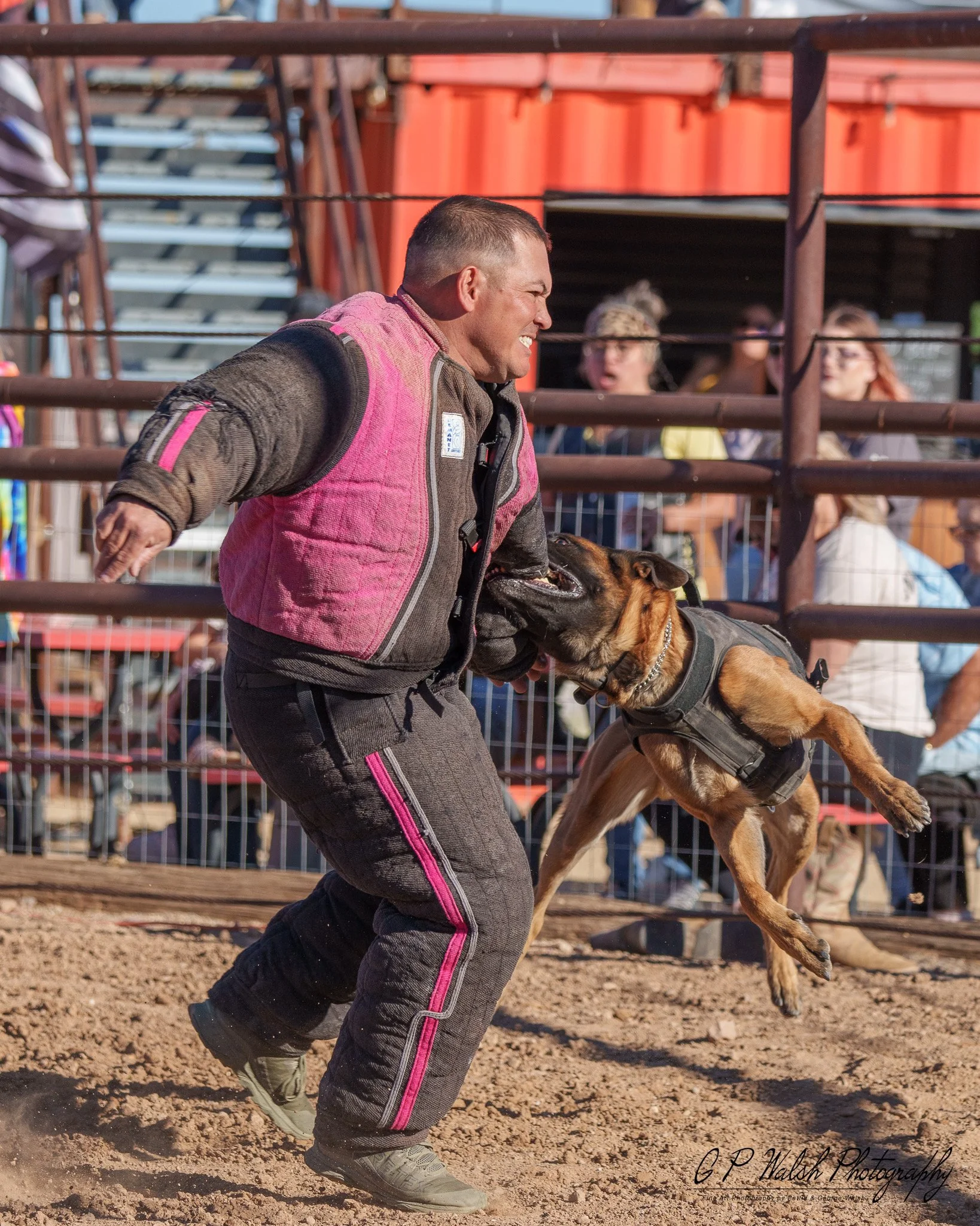 A man wearing a pink and black protective vest and gray sneakers is running with a dog in an outdoor arena. The man is smiling and holding the dog's leash, and the dog is leaping or running beside him. There are onlookers behind a metal fence, some w