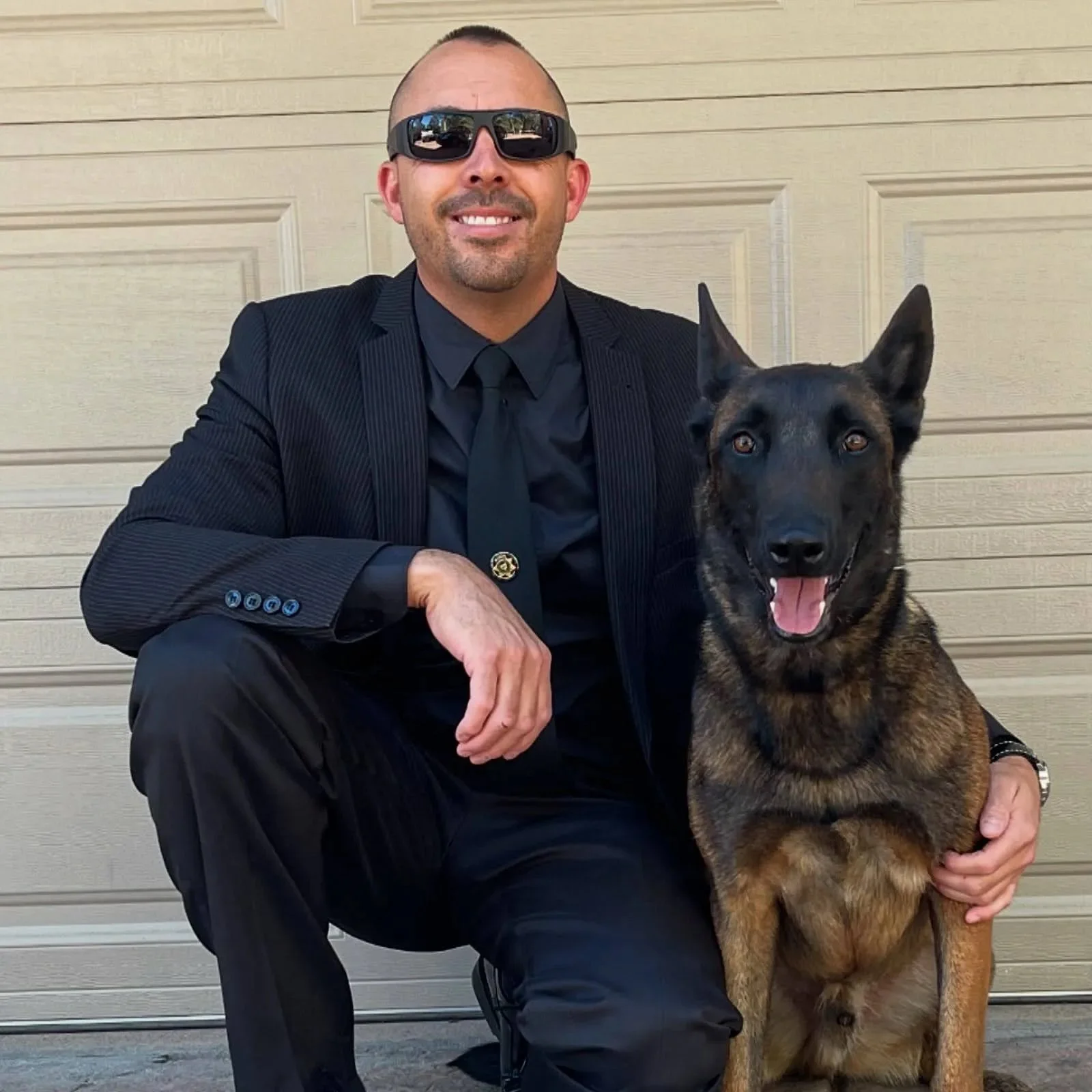A man dressed in a black suit and sunglasses, sitting beside a large German Shepherd dog with a black and tan coat, in front of a beige garage door.