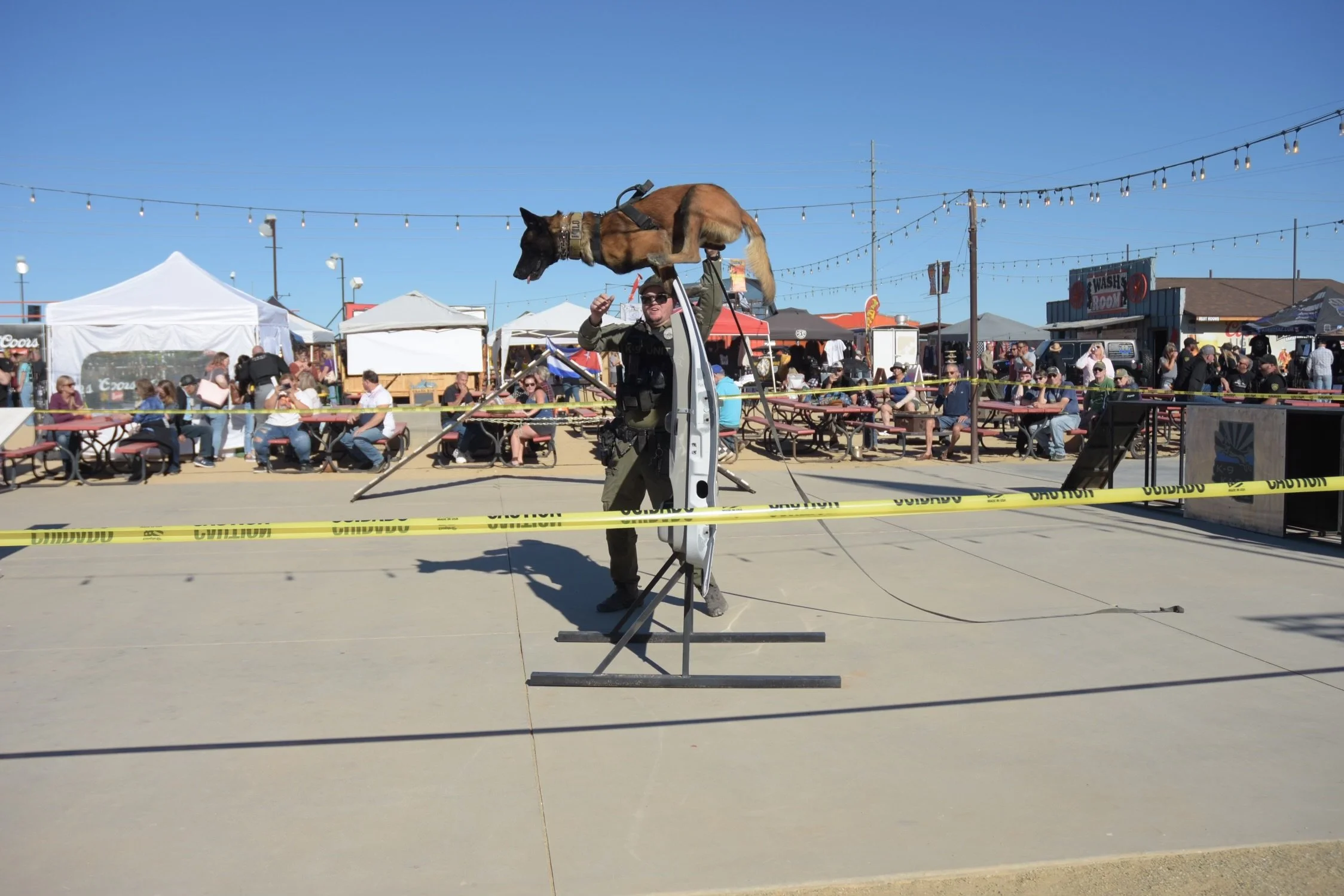 A police officer holding a large dog above his head during an outdoor event at a fair or festival, with people seated at picnic tables in the background. The scene is sunny with string lights overhead and temporary structures around.