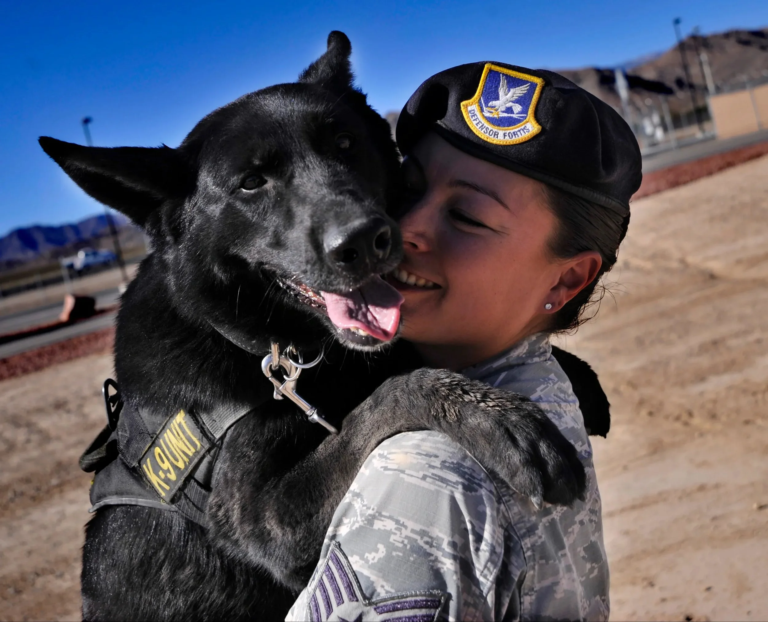 A woman in military uniform hugging a black police dog with a harness labeled 'K-9 UNIT' outdoors on a sunny day, with mountains in the background.