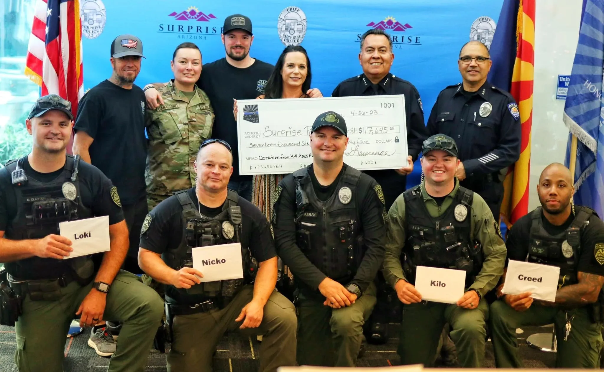 A group of police officers and civilians at an event, holding a large check dated April 20, 2023, with various signs indicating names like 'Loki,' 'Nicko,' 'Kilo,' 'Creed.' The background shows banners and flags, including the Arizona state flag, and a community or police department logo.