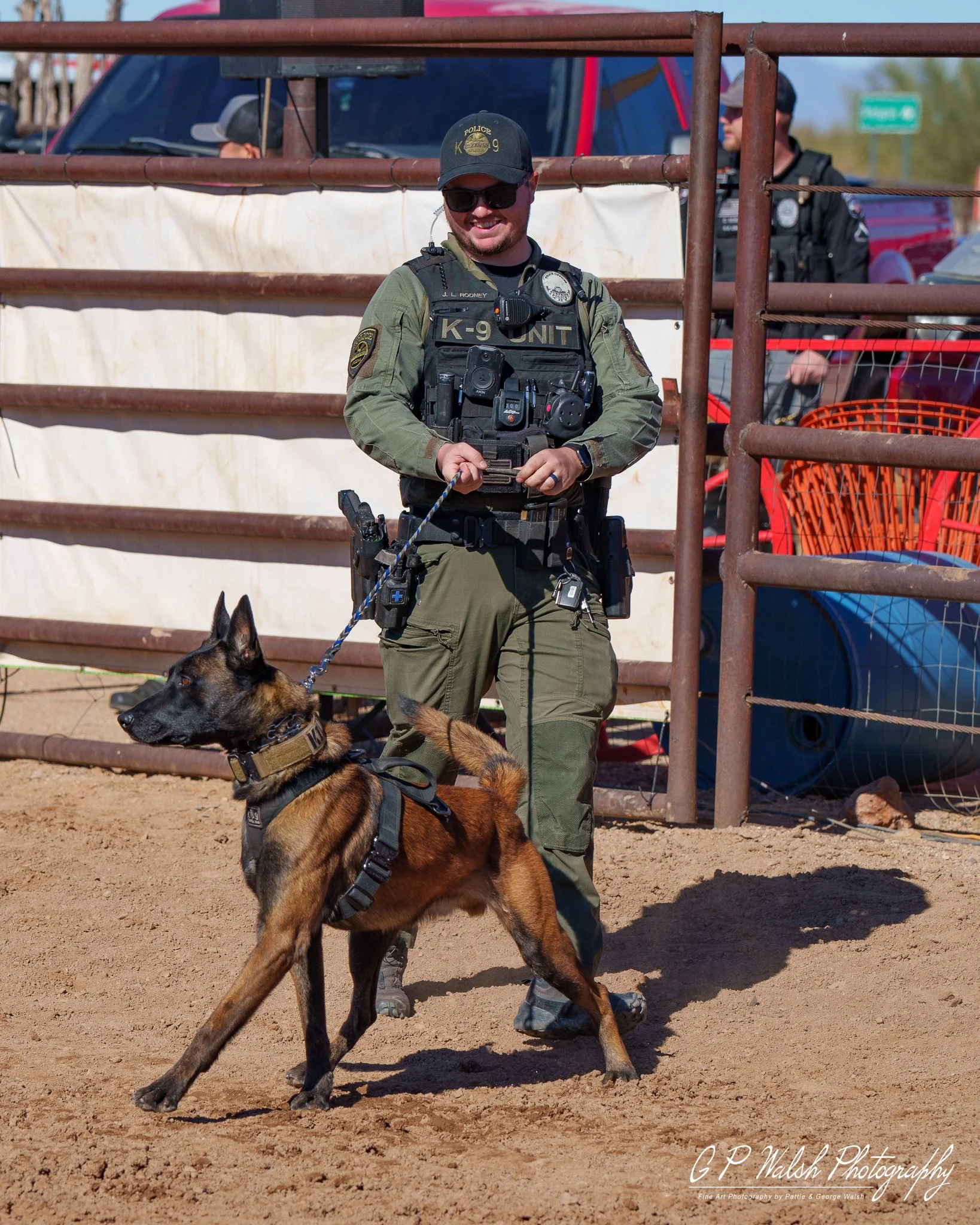 A police officer in tactical gear and sunglasses standing on dirt ground with a dog in the foreground. The dog is wearing a harness and looking to the side. Behind them are metal fences, a white wall, and a police vehicle with other officers in the b
