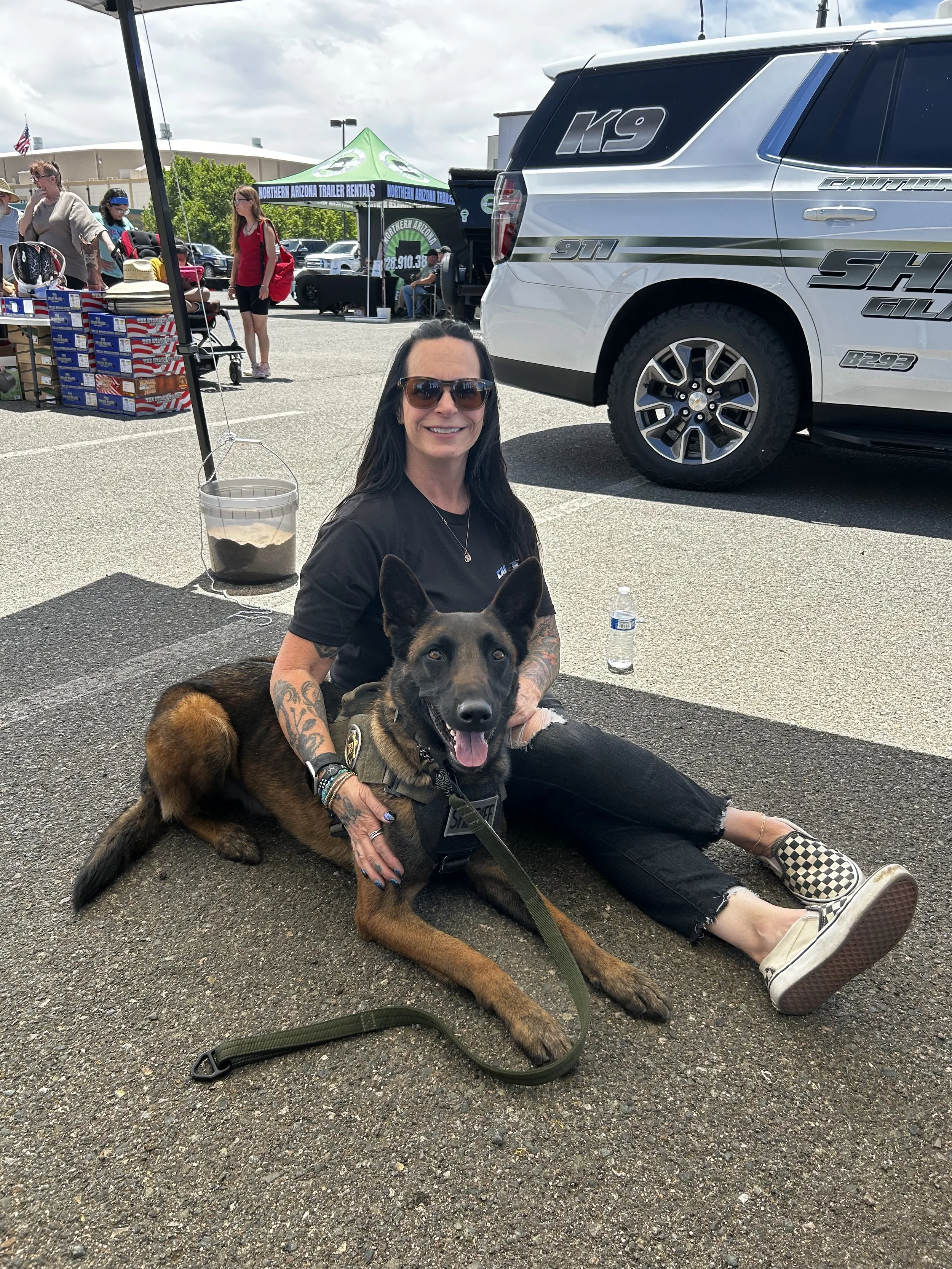 Woman with tattoos, wearing sunglasses, sitting on the parking lot ground, smiling, holding a German Shepherd dog, with a water bottle beside her, at an outdoor event with people, tents, and vehicles in the background.