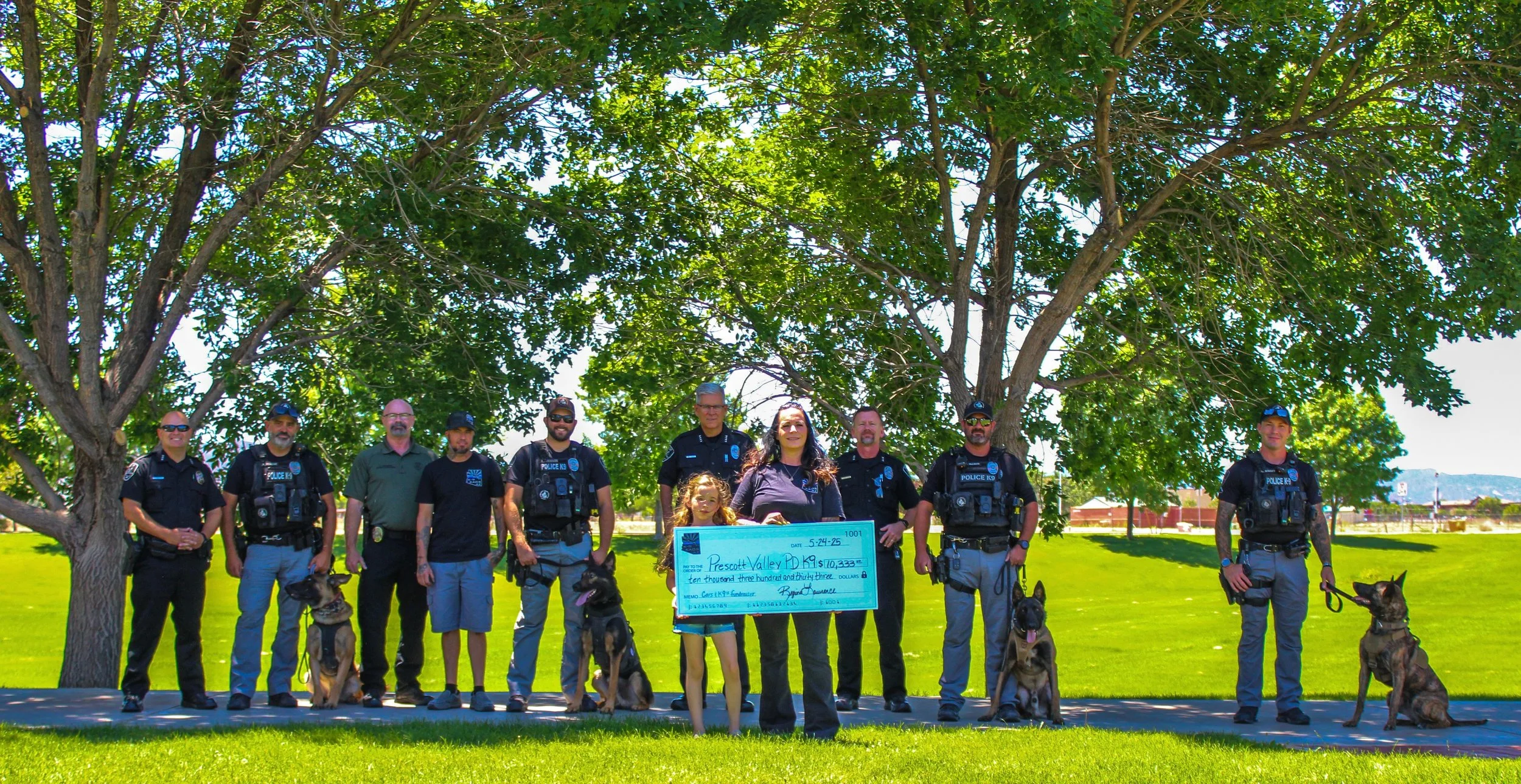Group of police officers and a woman with a girl holding a large check, standing under a large tree in a park on a sunny day.