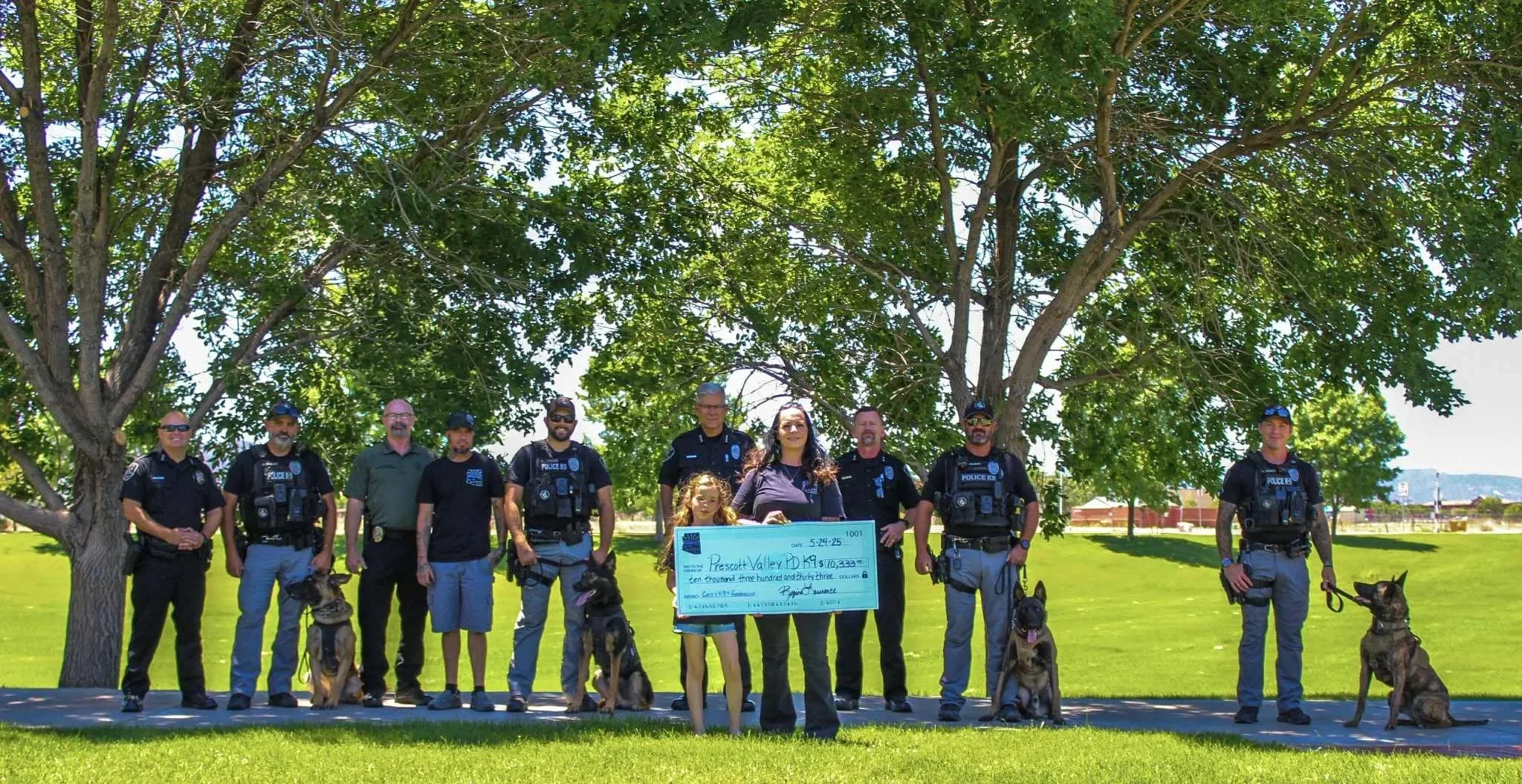 Group of police officers, a woman, and a young girl standing outdoors under a large tree on a sunny day, holding a large check for over ten thousand dollars while dogs sit beside them.