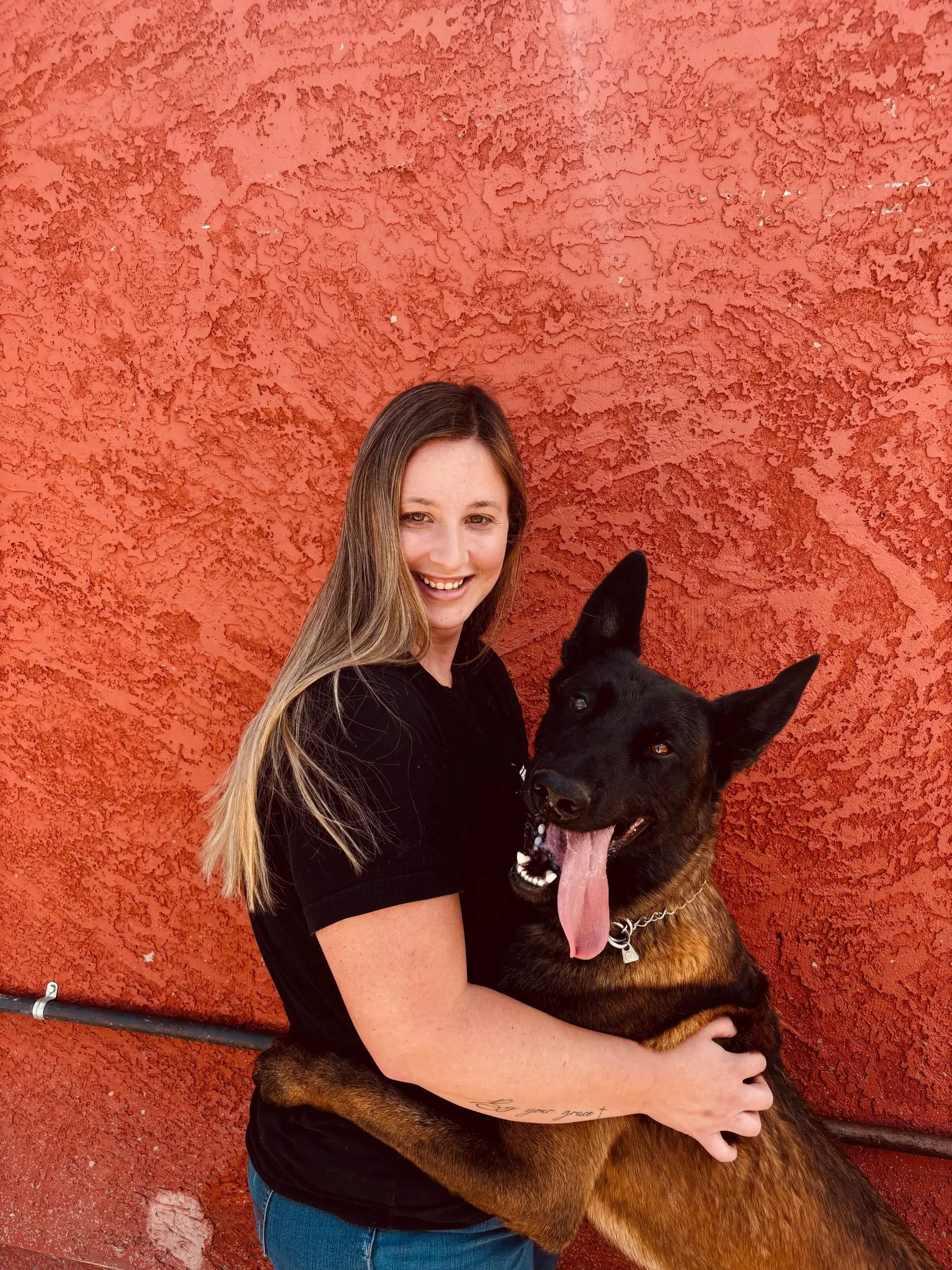 A woman with long brown hair smiling and hugging a large dog with black and brown fur, against an orange textured wall.
