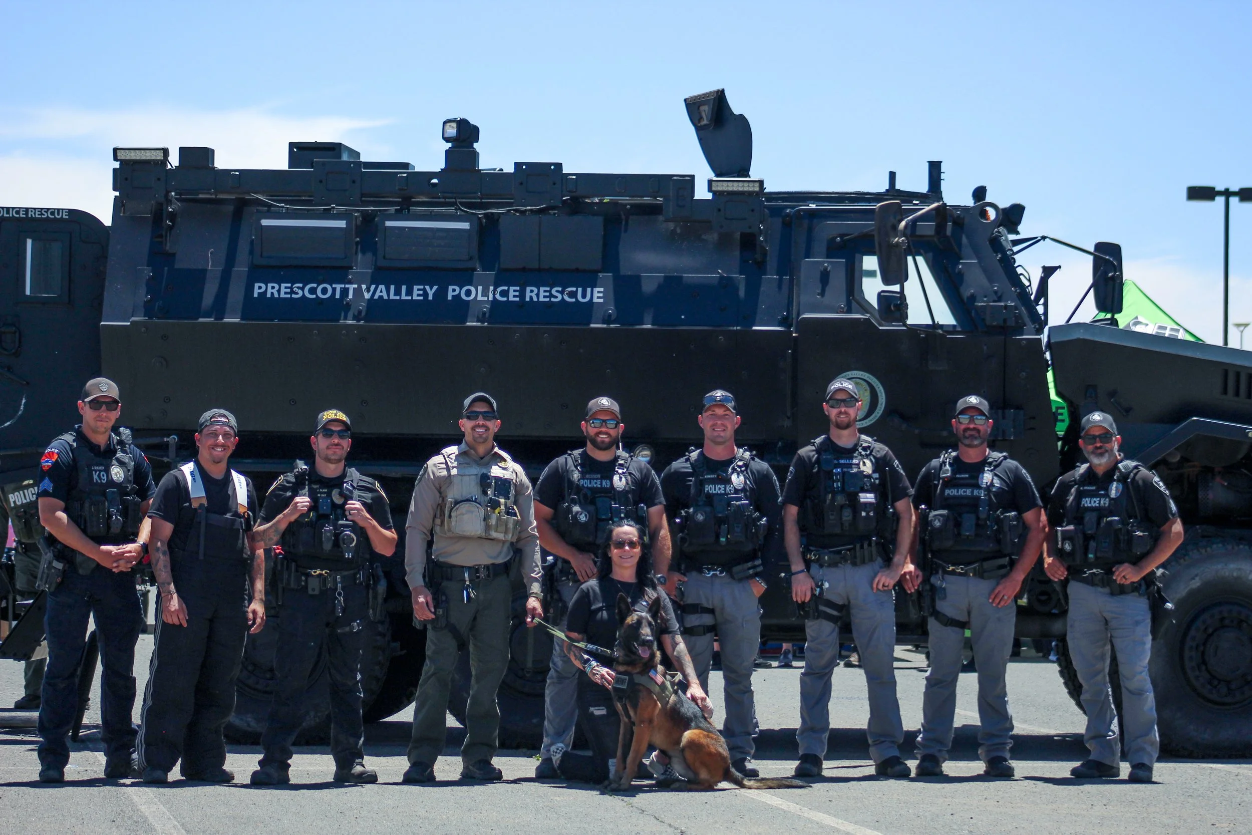A group of police officers, some in tactical gear and others in uniform, standing in front of a large police rescue vehicle labeled 'Prescott Valley Police Rescue,' with some officers accompanied by a police dog. The officers are smiling, and the scene appears to be at an outdoor event on a sunny day.