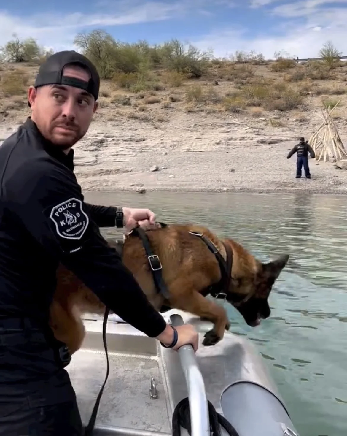 A police officer in uniform is holding a brown dog wearing a harness on a boat near a body of water, with a desert landscape and another person standing on the shore in the background.