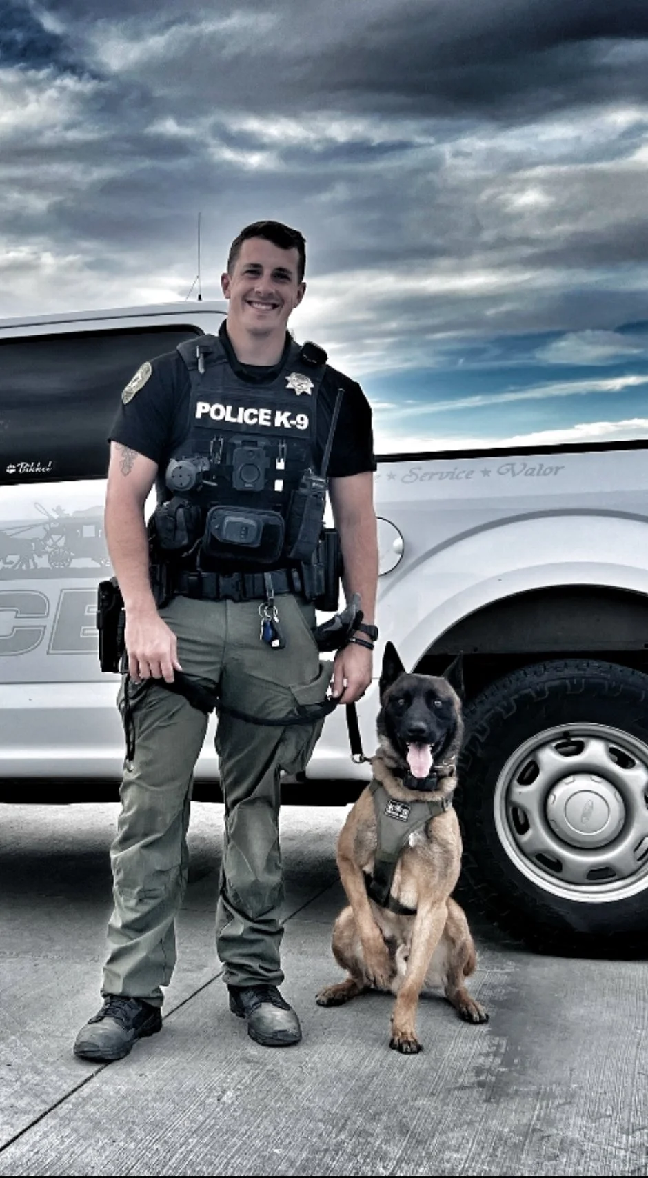 A police officer standing next to a police vehicle with his police dog, both wearing tactical gear, outdoors on a cloudy day.
