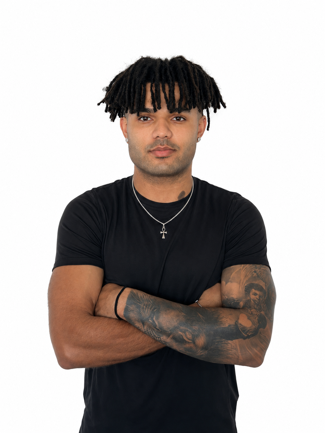 Portrait of a young man with dreadlocks, tattoos on arms, wearing a black t-shirt, silver necklace, and earrings, standing against a white background.
