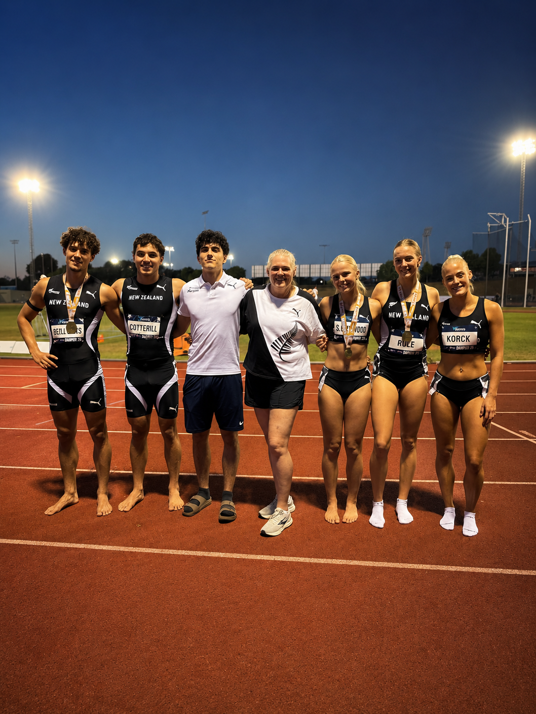 Group of seven athletes, five women and two men, standing on a track field at night. They are smiling and some are wearing medals. The women are in black sports bras and shorts with sneakers, and the men are in athletic shirts and shorts, some barefoot. They are posing together under stadium lights at an outdoor sports stadium.